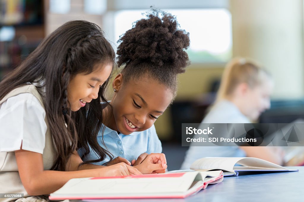 Happy female elementary age students in school uniforms are sitting at a desk in the school library and reading a book together. A Filipino elementary age girl sits with an African American classmate and reads a book together in the school library.
